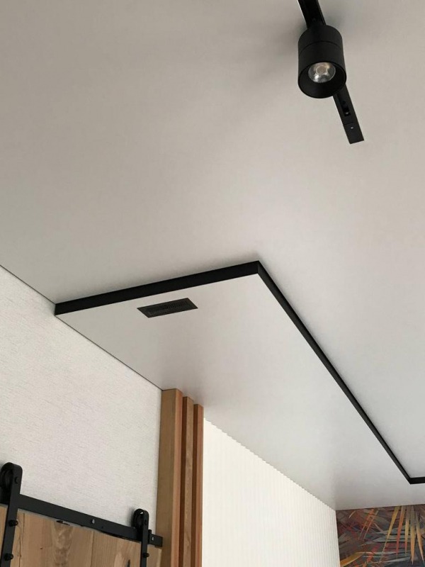 Fragment białego sufitu z czarną obwódką, oświetlenie szynowe z reflektorem, kratka wentylacyjna w suficie, dekoracyjna ściana z drewnianymi panelami i fragment drzwi przesuwnych.