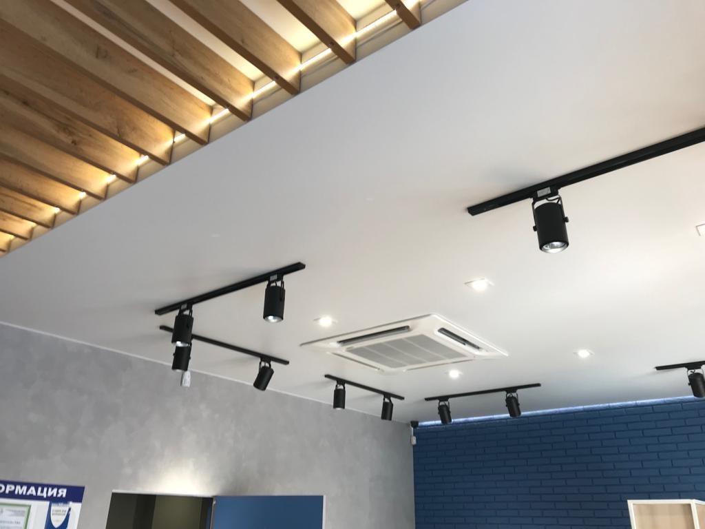 Biały sufit z zamontowanymi czarnymi szynami oświetleniowymi z reflektorami punktowymi, widoczna instalacja klimatyzacji i drewniane belki dekoracyjne
