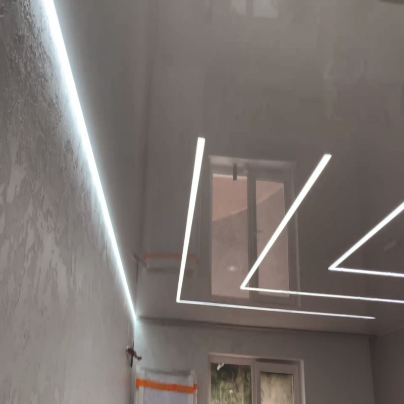 Biały, błyszczący sufit z wbudowanym oświetleniem LED w kształcie geometrycznym, widok z dołu, ściana z fakturą. Dwa okna w tle.