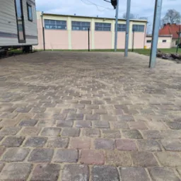 Nowa, ułożona kostka brukowa z piaskiem na spoinach, widok z niskiej perspektywy. W tle przyczepa kempingowa, budynki i ogrodzenie z siatki.