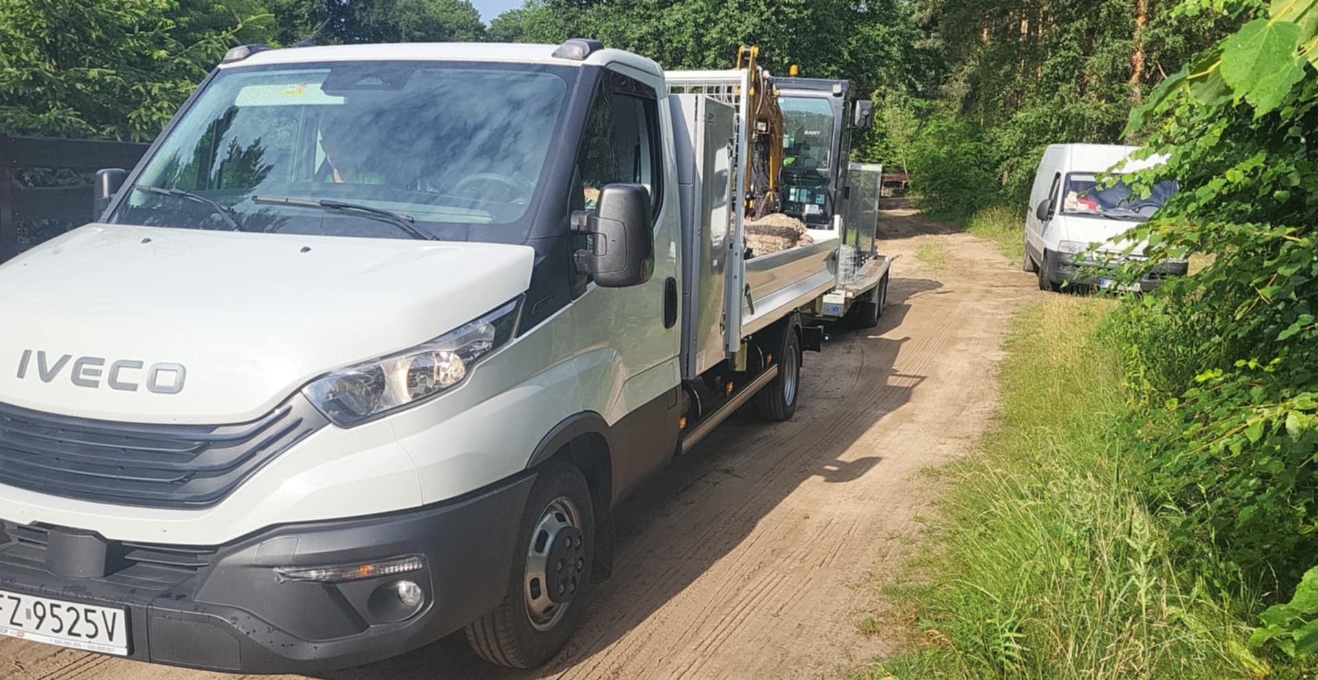 Biały samochód dostawczy Iveco z przyczepą, na której znajduje się mała koparka, na polnej drodze w otoczeniu zieleni. W tle widoczny drugi samochód dostawczy.