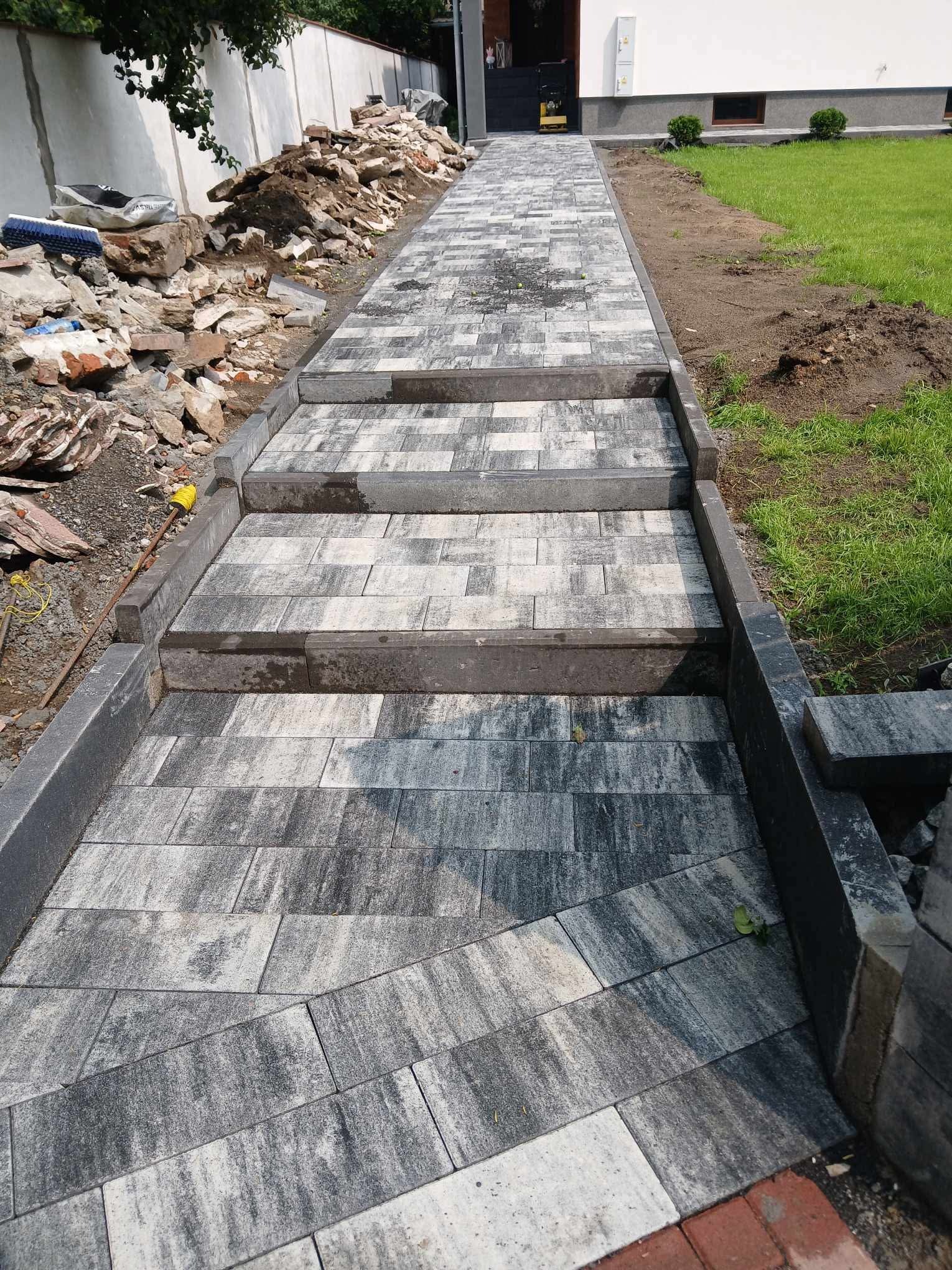 Ułożona ścieżka z kostki brukowej prowadząca do domu, z widocznymi schodami i fragmentem trawnika. Po lewej stronie hałda gruzu i narzędzia.