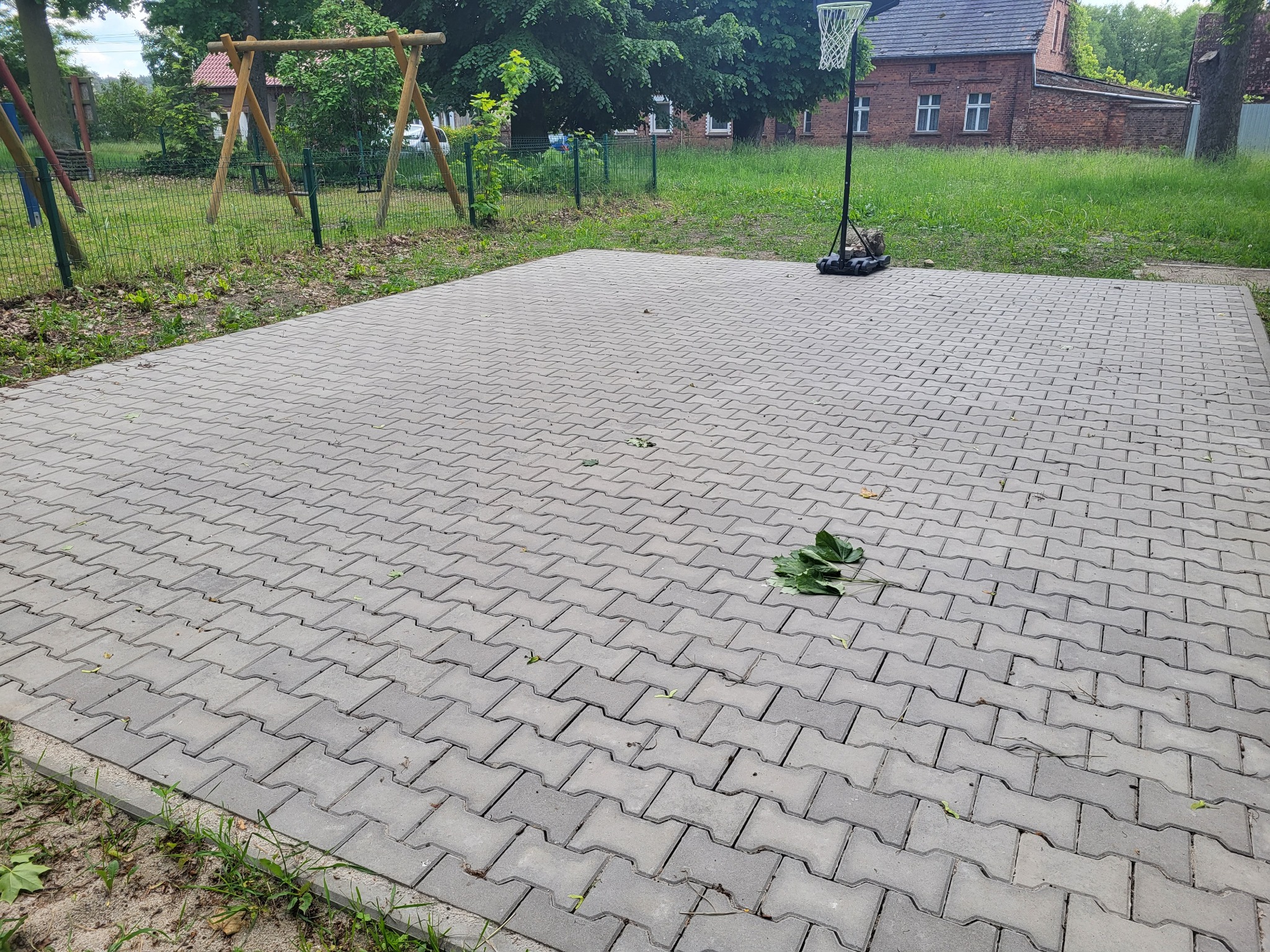 Szara kostka brukowa ułożona na placu zabaw z huśtawką i koszem do koszykówki w tle. Na kostce leżą zielone liście. Widok na budynek z czerwonej cegły.