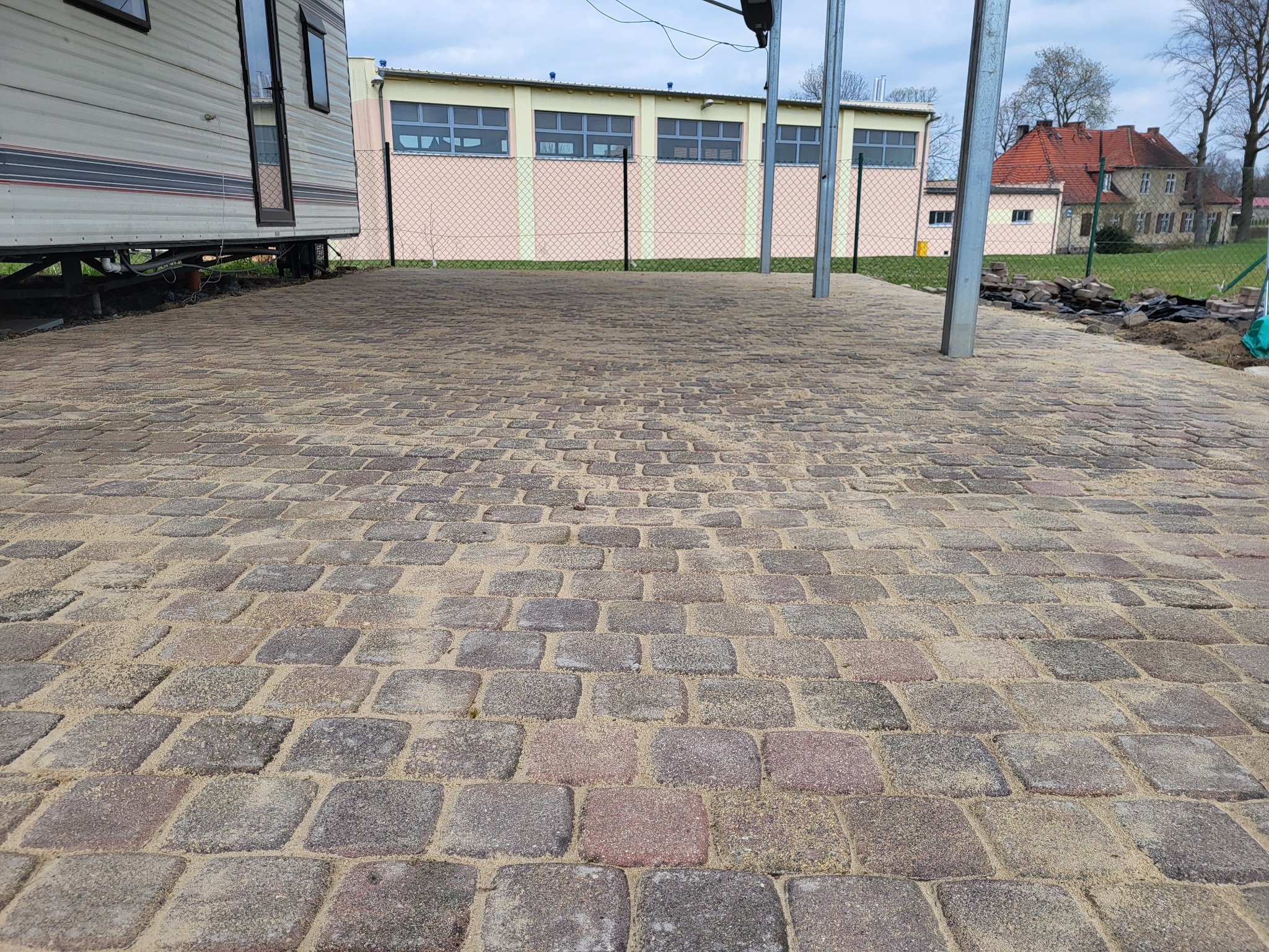 Nowa, ułożona kostka brukowa z piaskiem na spoinach, widok z niskiej perspektywy. W tle przyczepa kempingowa, budynki i ogrodzenie z siatki.