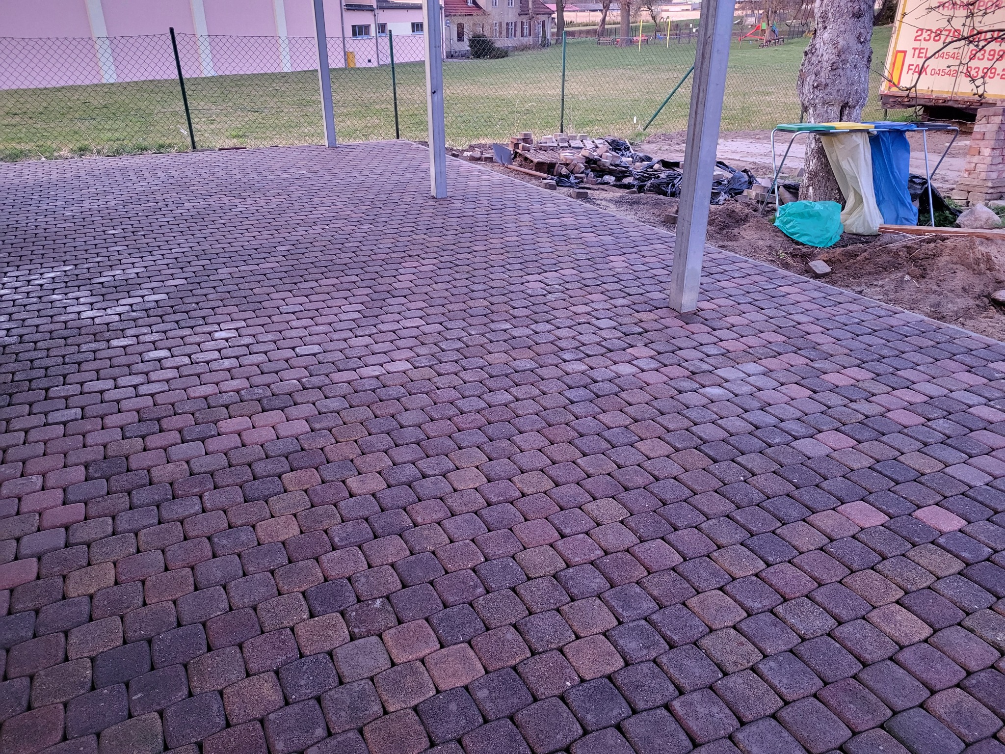Ukończona nawierzchnia z kostki brukowej w odcieniach fioletu i brązu, z widocznymi słupami zadaszenia i przygotowanym terenem pod dalsze prace.