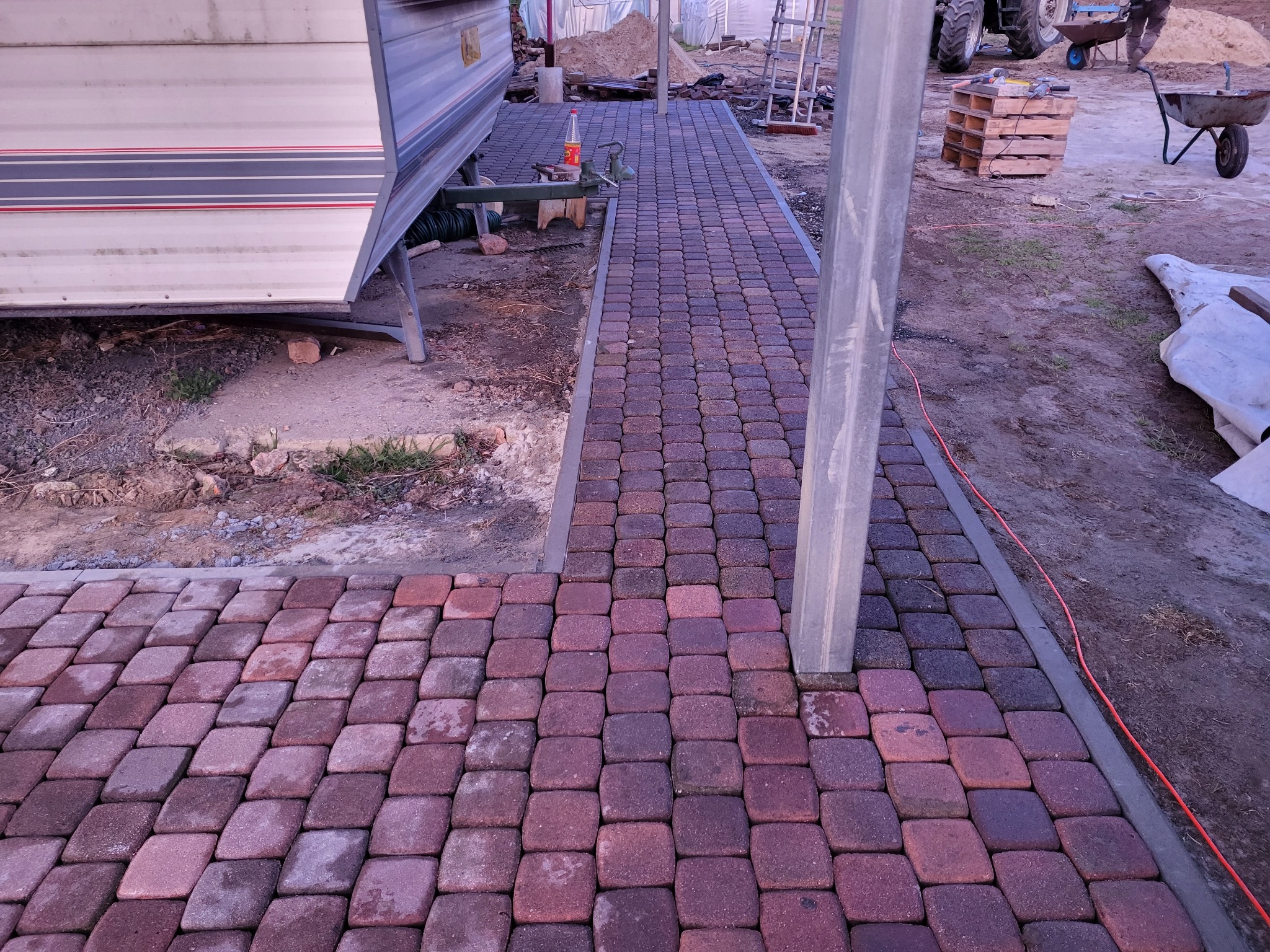 Ułożona, czerwono-brązowa kostka brukowa tworząca ścieżkę obok przyczepy kempingowej i elementów budowlanych, sugerująca prace brukarskie w toku.