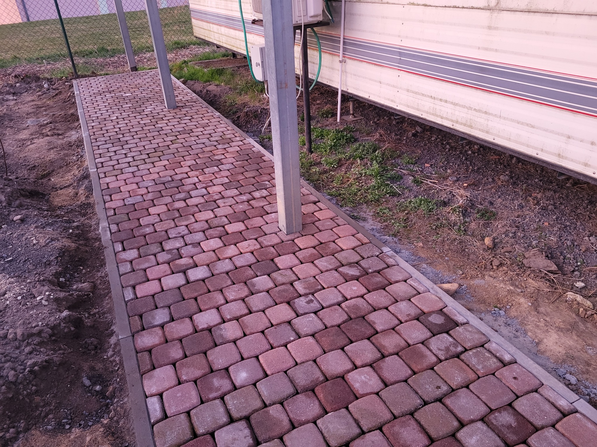 Ułożona kostka brukowa w odcieniach czerwieni i brązu, obramowana szarym obrzeżem, prowadząca wzdłuż przyczepy kempingowej. Słupy podtrzymujące zadaszenie.