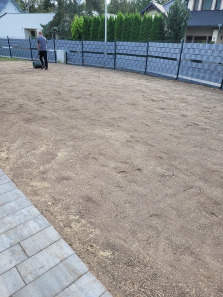 Przygotowany teren pod trawnik z widocznym wałem i fragmentem kostki brukowej. W tle szary płot panelowy i zabudowa jednorodzinna.