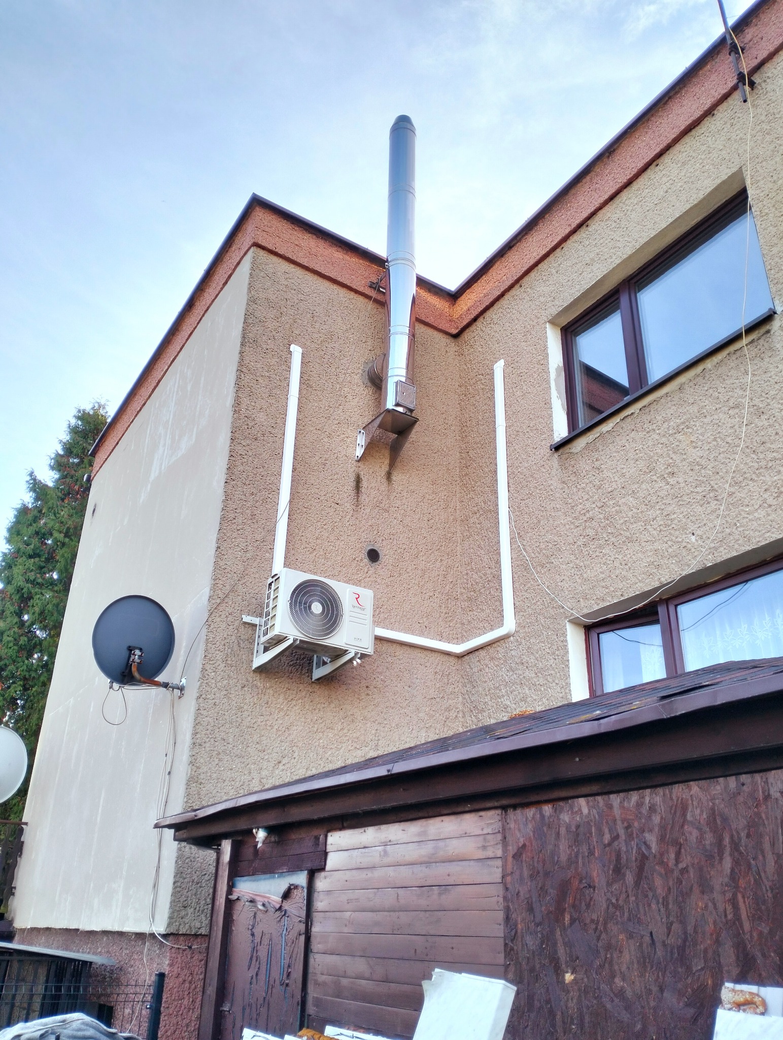 Zewnętrzna jednostka klimatyzacji zamontowana na elewacji domu z widocznymi rurami odprowadzającymi, obok zainstalowana antena satelitarna, widok z dołu.