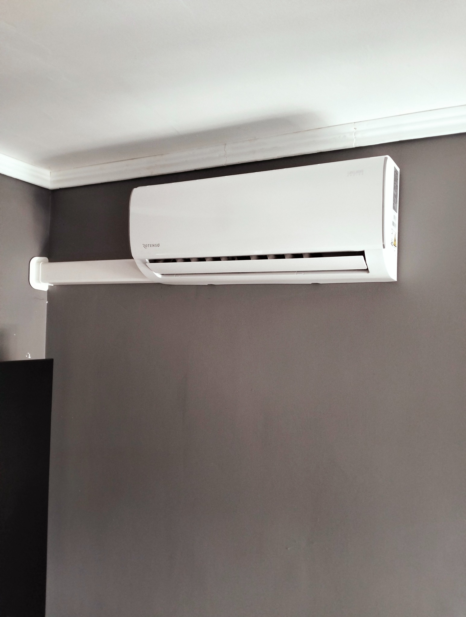 Biała jednostka klimatyzacyjna Rotenso zamontowana na szarej ścianie w pomieszczeniu, widoczna instalacja odprowadzająca skropliny.