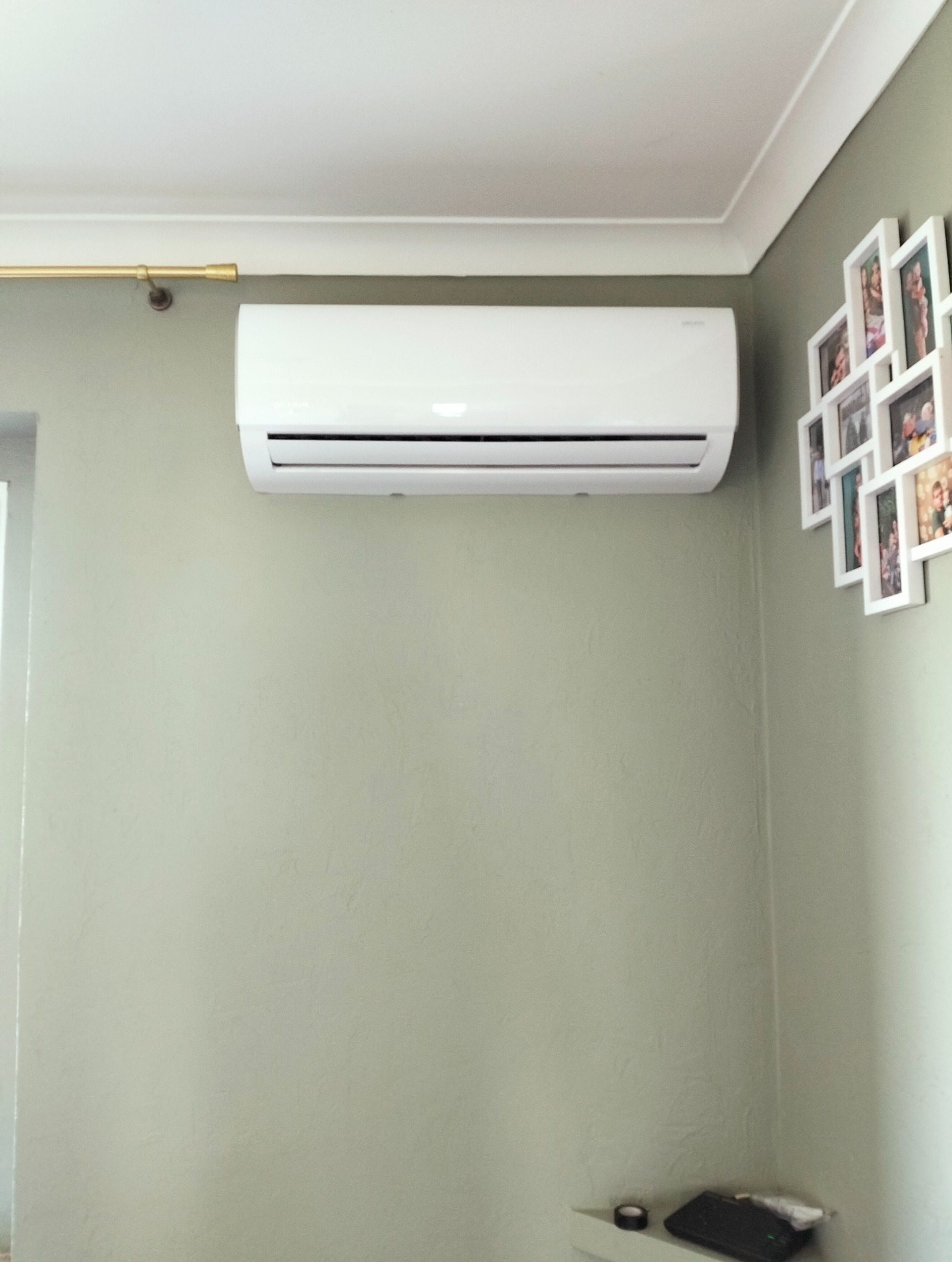 Biała jednostka klimatyzacji na ścianie w pokoju z zielonymi ścianami i ramkami ze zdjęciami.