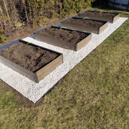 GreeenGarden Mateusz Kwidzinski - Cztery podwyższone grządki z brązowego drewna wypełnione ziemią, ustawione na białym żwirze, z młodymi sadzonkami, na tle zielonego żywopłotu i trawnika.