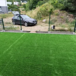 Świeżo ułożona, intensywnie zielona trawa syntetyczna w ogrodzie, otoczona betonową obwódką i siatką ogrodzeniową. W tle zaparkowany samochód osobowy.