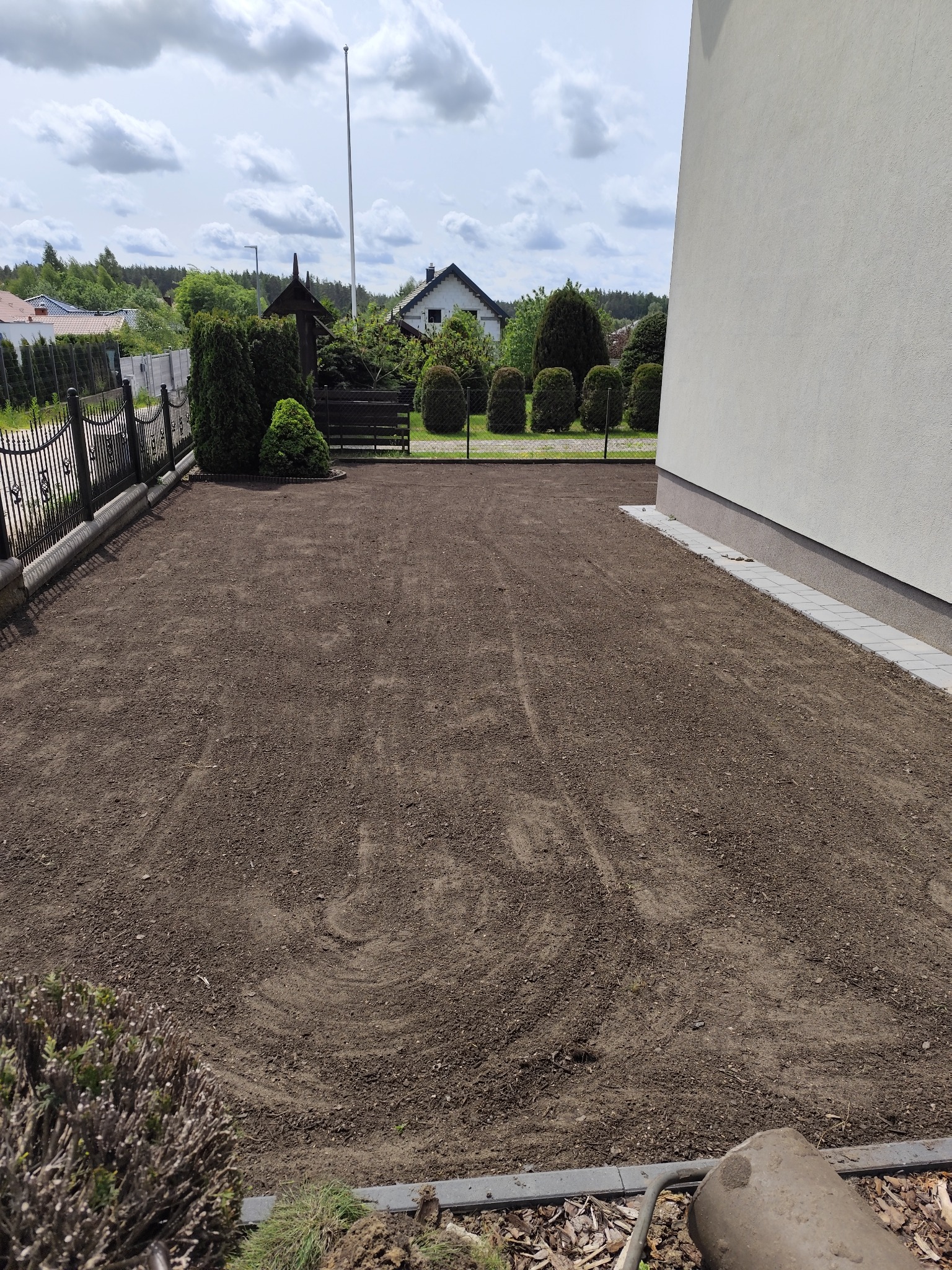 Przygotowany teren pod trawnik przy domu jednorodzinnym, widoczne ślady po walcu, obrzeża z kostki, w tle krzewy i drzewa. Słoneczny dzień.