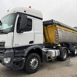TRANS-TECH PIŁA SPÓŁKA Z OGRANICZONĄ ODPOWIEDZIALNOŚCIĄ - Ciężarówka Mercedes-Benz Actros 1843 z żółtą naczepą wywrotką, częściowo przykrytą plandeką, zaparkowana na brukowanej powierzchni, widok z boku na pochmurnym tle.