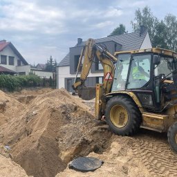 TRANS-TECH PIŁA SPÓŁKA Z OGRANICZONĄ ODPOWIEDZIALNOŚCIĄ - Żółta koparko-ładowarka 428D przy wykopie w piaszczystej ziemi na tle domów jednorodzinnych, widoczna czarna rura drenażowa i właz kanalizacyjny.