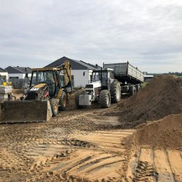 TRANS-TECH PIŁA SPÓŁKA Z OGRANICZONĄ ODPOWIEDZIALNOŚCIĄ - Żółta koparko-ładowarka CAT i biały ciągnik z przyczepą na placu budowy, w tle budynki mieszkalne z ciemnymi dachami, widoczny wykopany piasek i ślady opon.