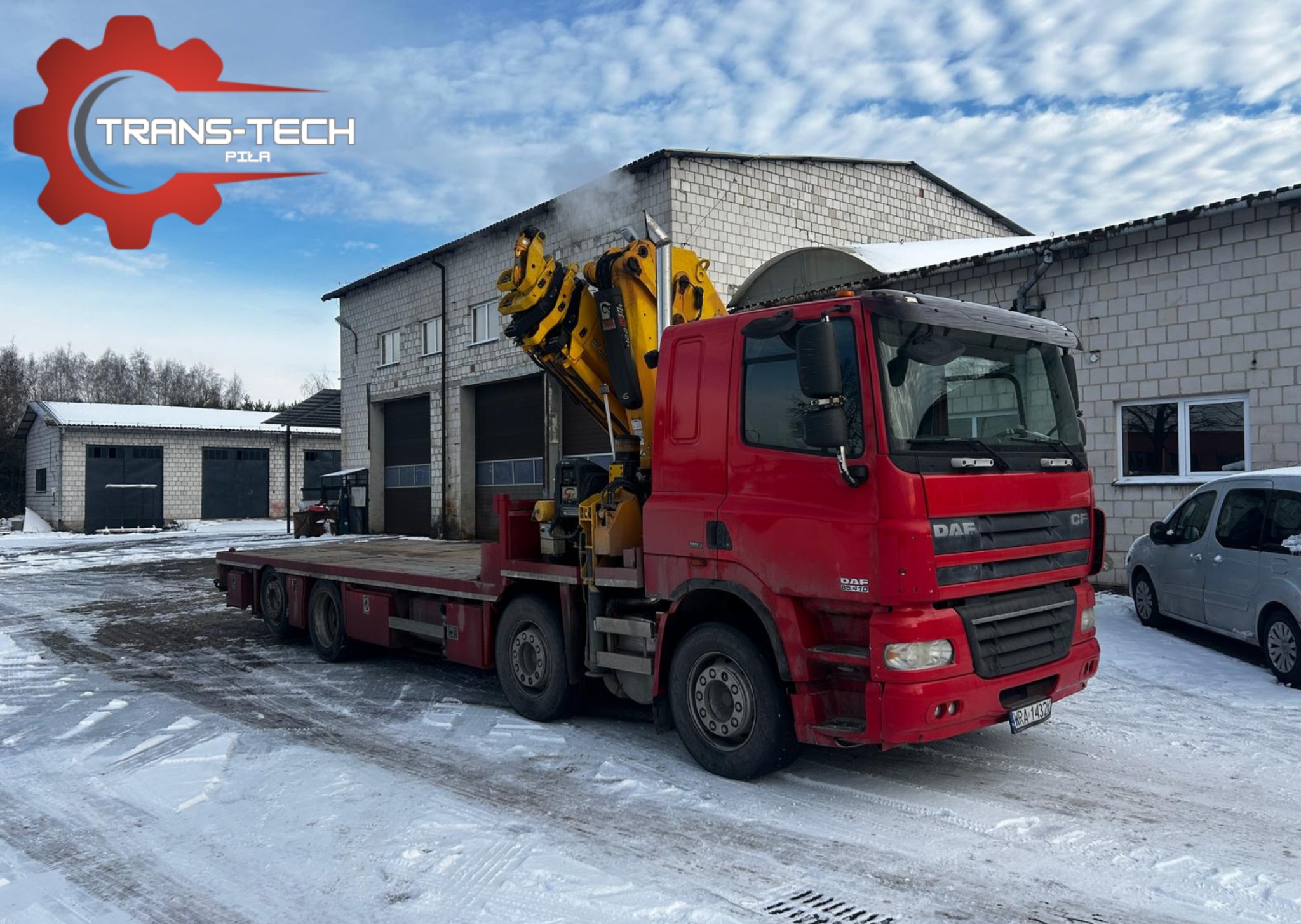Czerwona ciężarówka DAF z żółtym dźwigiem HDS na platformie, zaparkowana na śniegu przed budynkiem magazynowym z cegły.