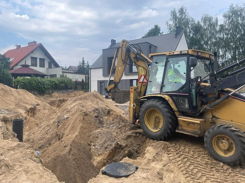 Żółta koparko-ładowarka 428D przy wykopie w piaszczystej ziemi na tle domów jednorodzinnych, widoczna czarna rura drenażowa i właz kanalizacyjny.