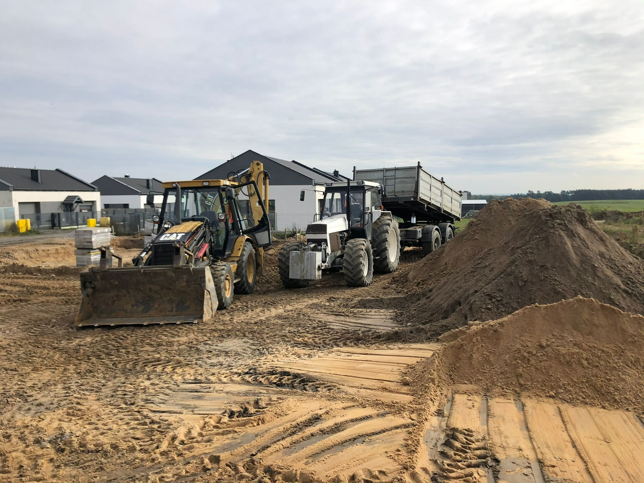Żółta koparko-ładowarka CAT i biały ciągnik z przyczepą na placu budowy, w tle budynki mieszkalne z ciemnymi dachami, widoczny wykopany piasek i ślady opon.