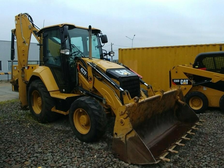 Żółta koparko-ładowarka Caterpillar 427F zaparkowana na żwirowym podłożu, obok żółtego kontenera i mniejszej ładowarki Caterpillar 236D.