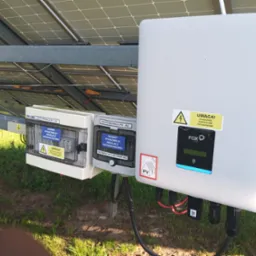 Instalacja fotowoltaiczna: falownik FOX ESS, rozdzielnice PV-DC i AC. Widoczne panele słoneczne, okablowanie i etykiety ostrzegawcze. System fotowoltaiczny w otoczeniu zieleni.