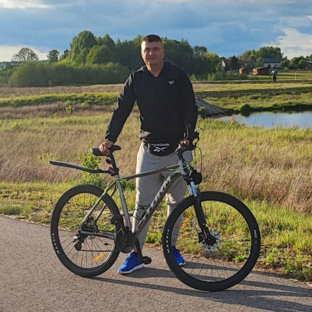 Mężczyzna w sportowym stroju stoi obok roweru górskiego na ścieżce, w tle łąka, zbiornik wodny i zabudowania. Pochmurne niebo nadaje scenerii stonowany charakter.