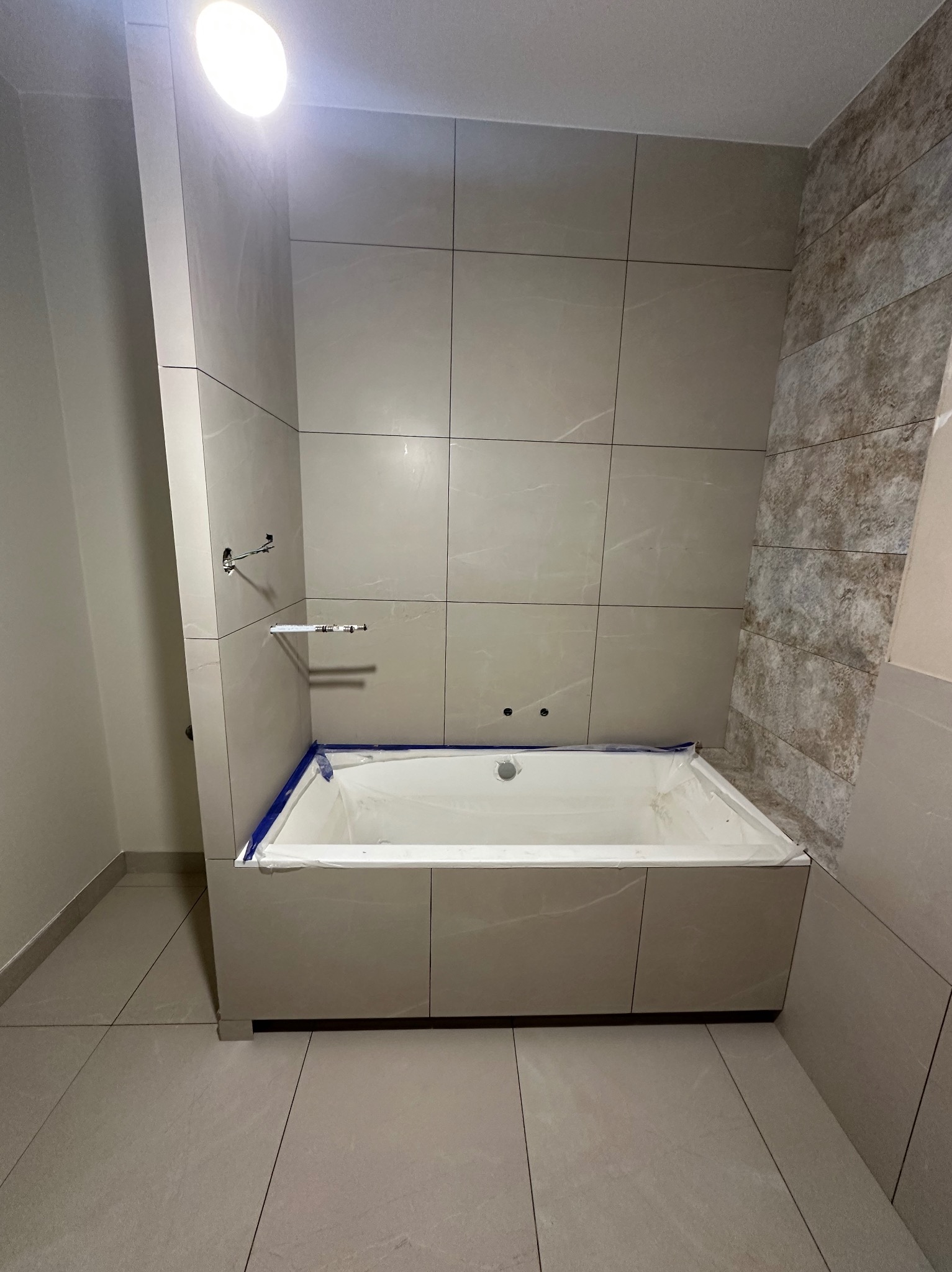 Wykończona łazienka: beżowe płytki na ścianach i obudowie wanny, widoczne przyłącza armatury, zabezpieczona wanna. Stonowana kolorystyka, nowoczesny design.