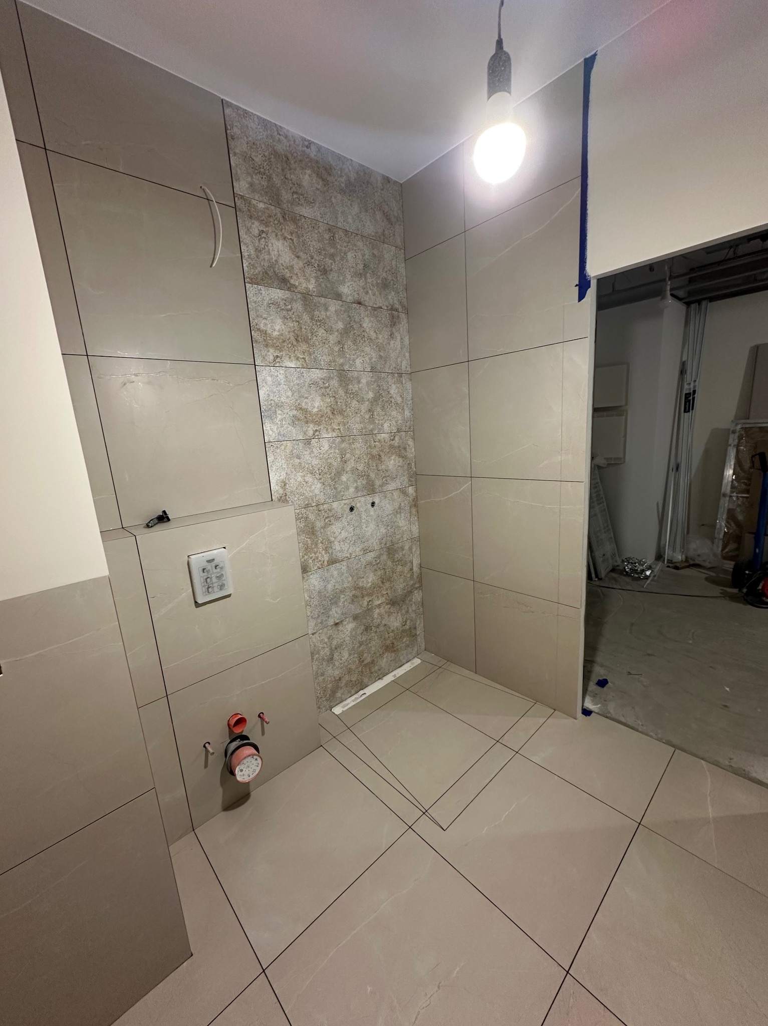 Beżowe płytki imitujące kamień w łazience, z elementami dekoracyjnymi. Widoczne przygotowane przyłącza wodno-kanalizacyjne i fragment otwartego przejścia do innego pomieszczenia.