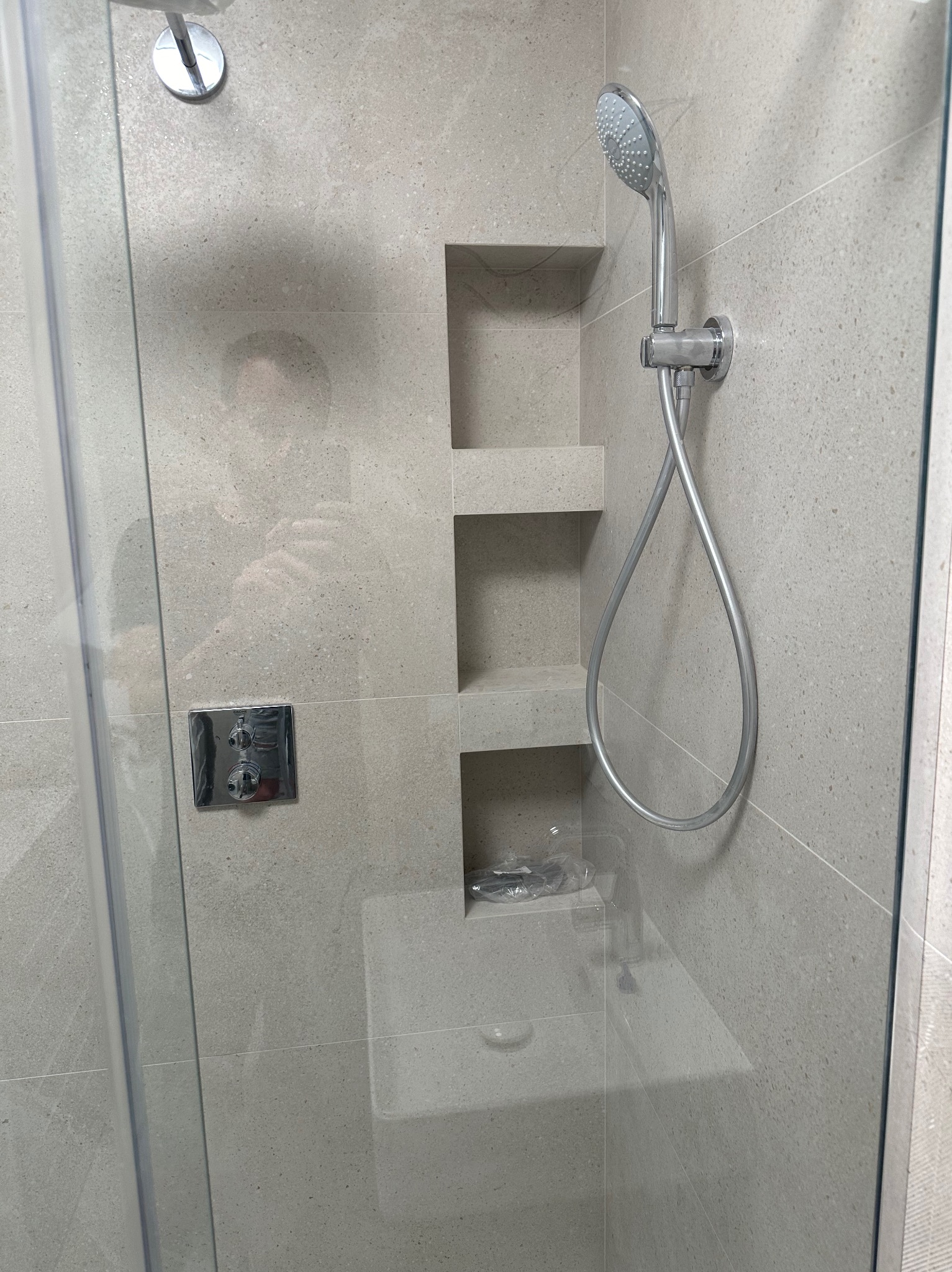 Nowoczesna kabina prysznicowa z szarymi płytkami, wnękami na kosmetyki i deszczownicą. Minimalistyczny design, szklane drzwi, chromowana armatura.