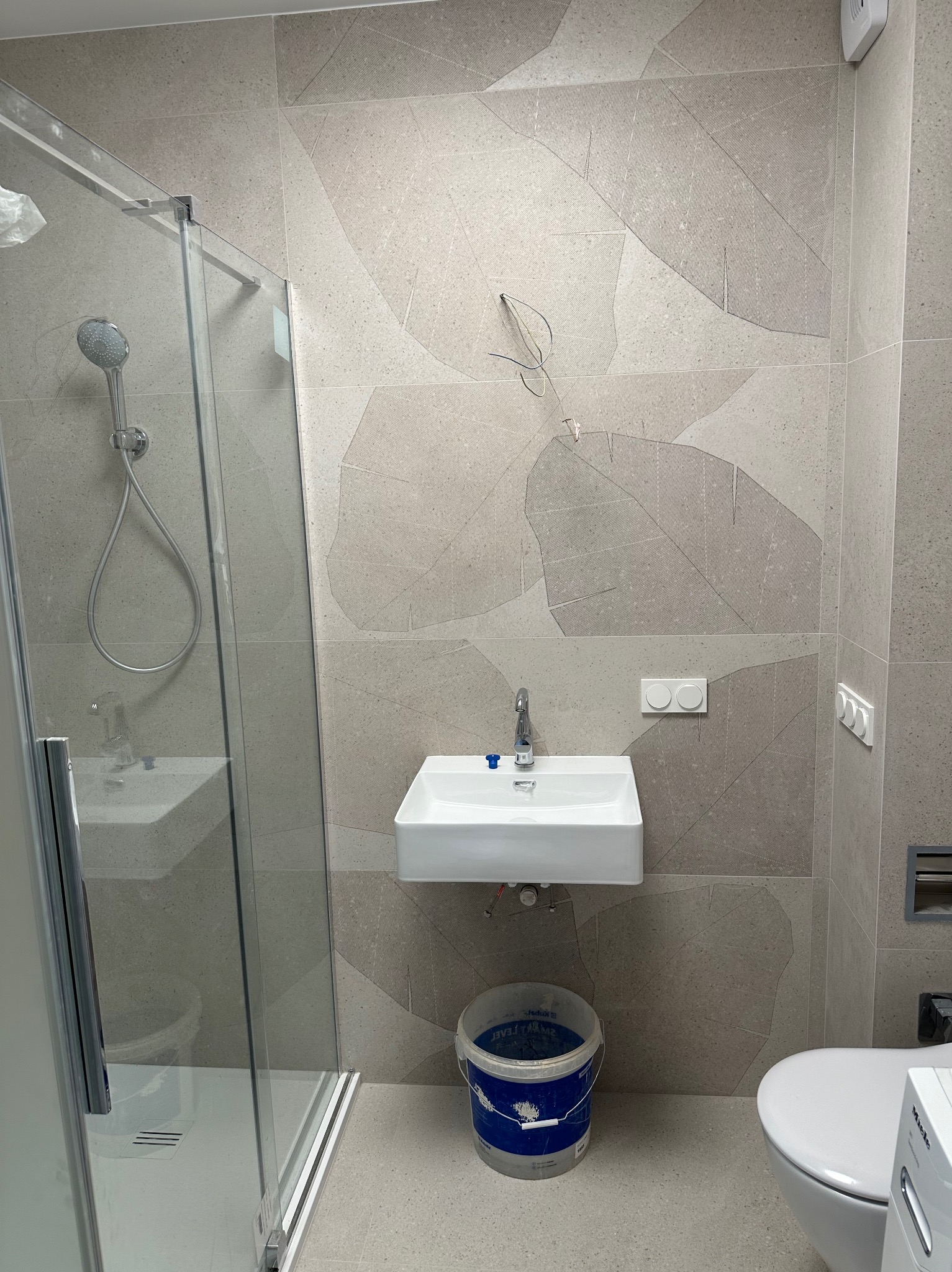 Wykończona łazienka w szarościach: kabina prysznicowa, umywalka nablatowa, toaleta. Na ścianach płytki z delikatnym wzorem liści. Widoczny kubeł malarski.