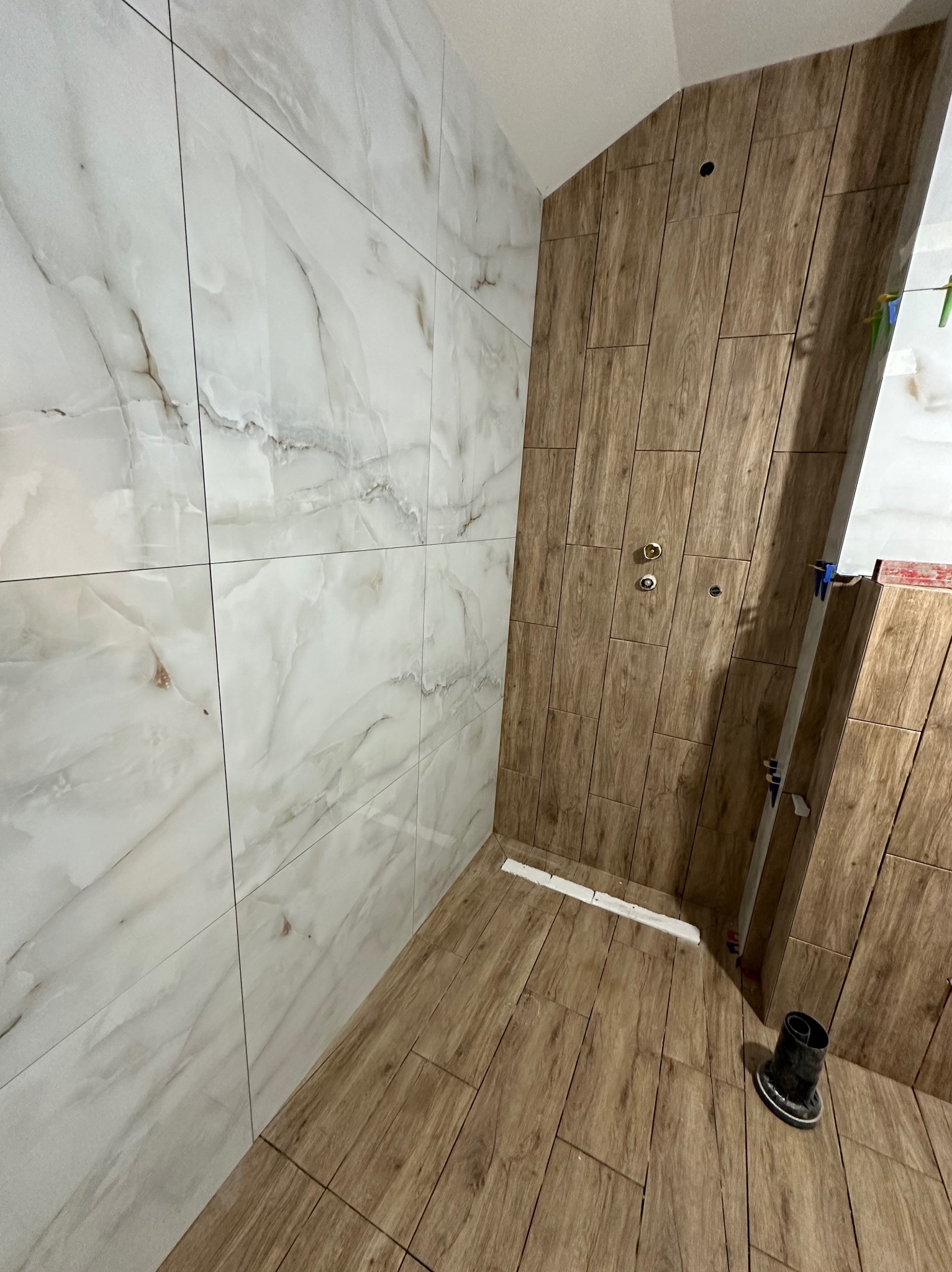 Wykończona kabina prysznicowa z połączeniem płytek imitujących marmur i drewno. Widoczny odpływ liniowy i elementy instalacji.