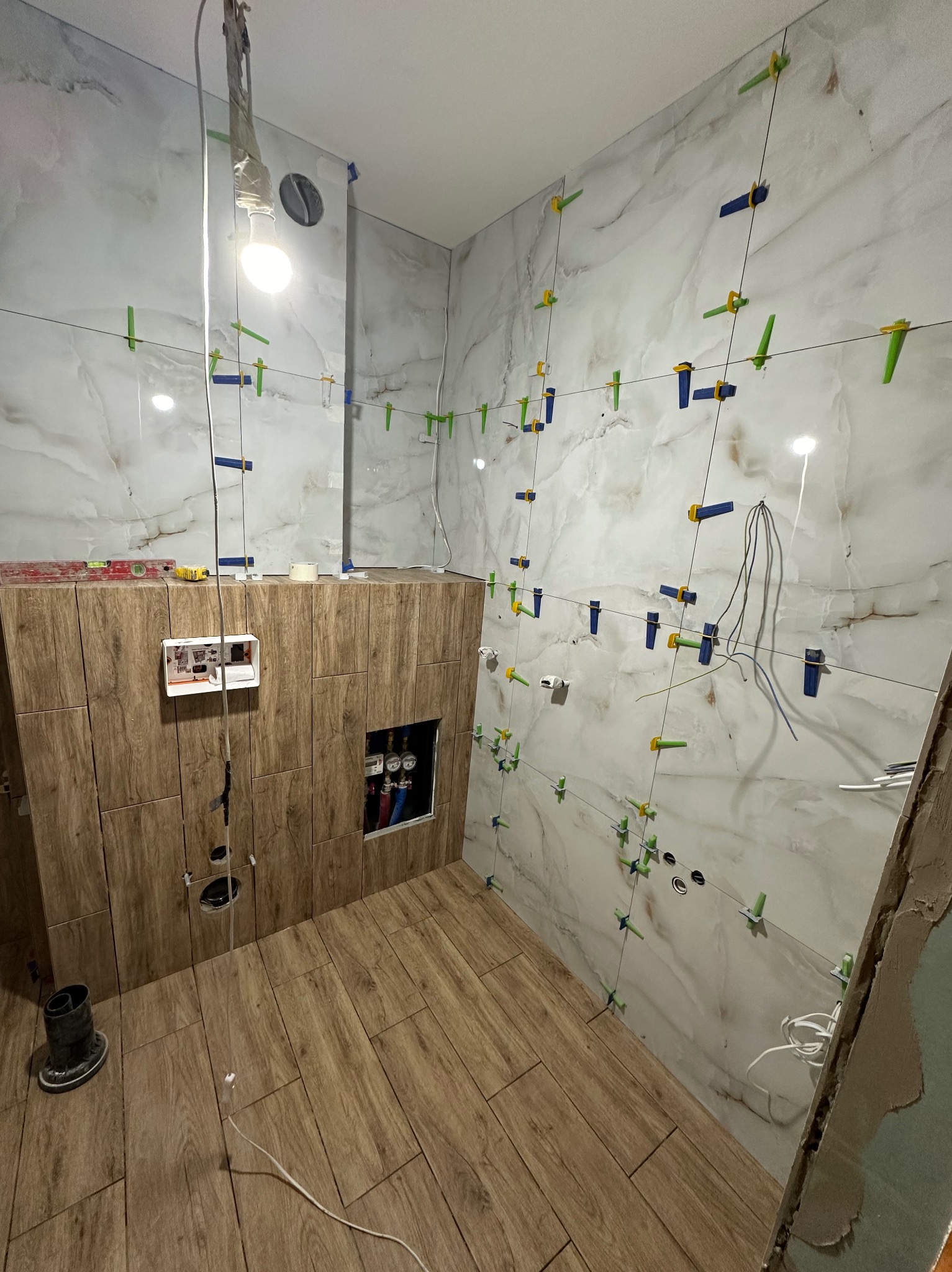 Łazienka w trakcie wykańczania: marmurowe płytki na ścianach z systemem poziomowania, drewnopodobna podłoga, widoczne instalacje i narzędzia.