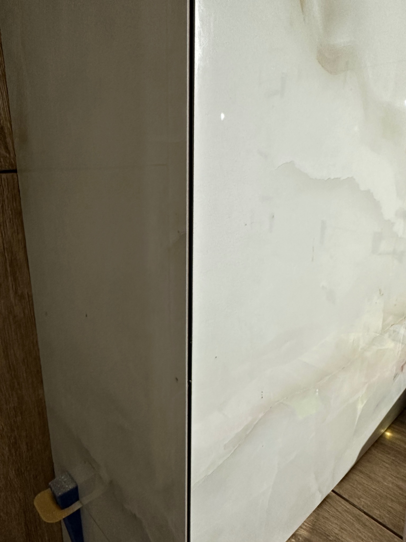 Białe płytki imitujące marmur na ścianie, z widocznym krzyżykiem glazurniczym i fragmentem drewnopodobnej podłogi, ukazujące wykończenie łazienki.