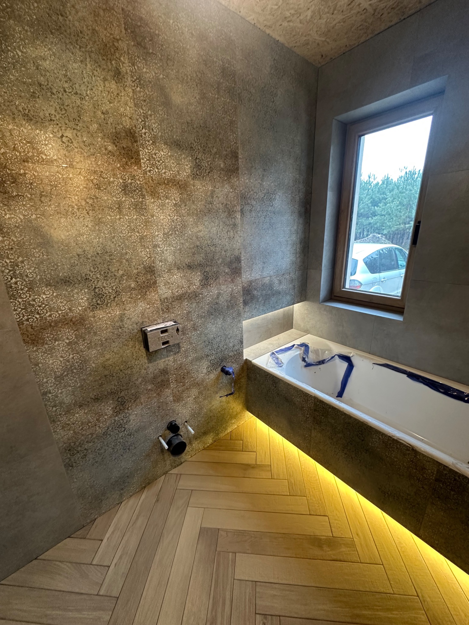 Wykończona łazienka z wanną zabezpieczoną folią, podświetleniem LED i wzorzystymi płytkami. Widoczny fragment okna i ułożona w jodełkę podłoga.