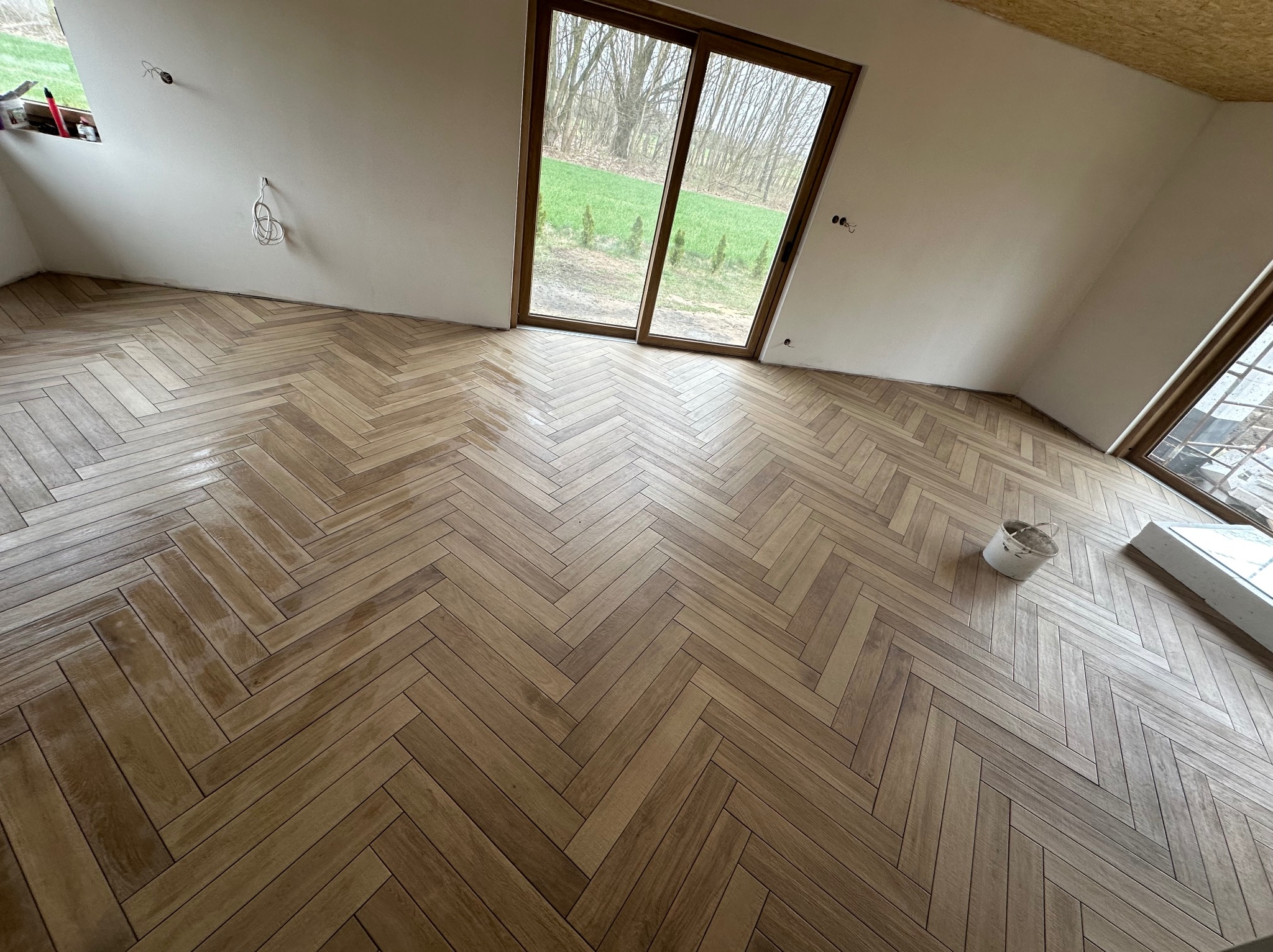 Parkiet ułożony we wzór jodełki w jasnym pokoju z dużym oknem balkonowym. Widoczne niedoskonałości ścian i wystające kable. Wiadro na podłodze.