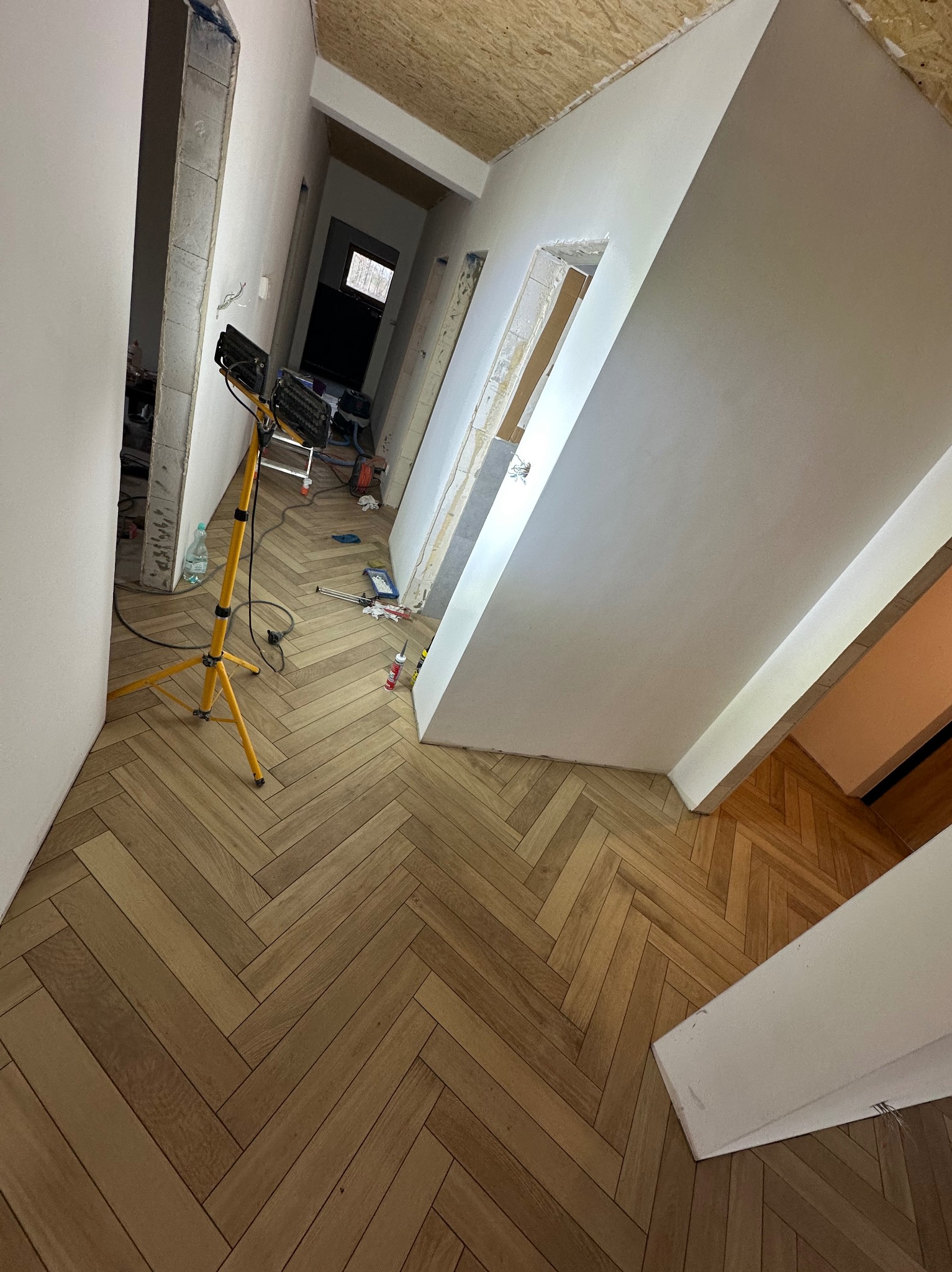 Układanie parkietu w jodełkę w korytarzu, widoczna lampa budowlana i narzędzia. Ściany w trakcie wykańczania, sufit z płyt OSB.