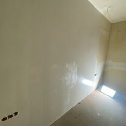 PROEFEKT Michał Czaplicki - Świeżo położona gładź szpachlowa na ścianie w rogu pokoju, widoczne puszki elektryczne bez osprzętu, oświetlenie budowlane na podłodze, ślady po pracach budowlanych na podłodze.