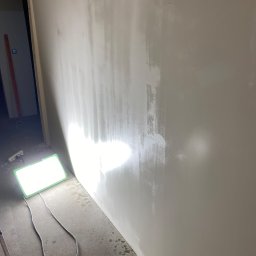 PROEFEKT Michał Czaplicki - Ściana w trakcie nakładania warstwy wygładzającej, oświetlona halogenem budowlanym, widoczne nierówności i ślady po szpachli.