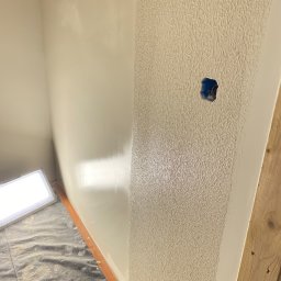 PROEFEKT Michał Czaplicki - Fragment wnętrza w trakcie prac wykończeniowych, widoczna ściana z nałożoną strukturą dekoracyjną obok gładkiej, pomalowanej ściany. Na podłodze folia ochronna i lampa budowlana. Widoczne puszki...