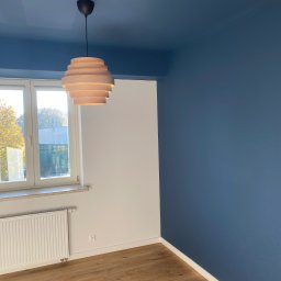 PROEFEKT Michał Czaplicki - Pomieszczenie po malowaniu z granatowym sufitem i ścianą, drewnianą podłogą, białym grzejnikiem i lampą z drewnianych kręgów.