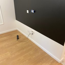 PROEFEKT Michał Czaplicki - Pokój po malowaniu z dwukolorową ścianą: czarny pas na górze i biały na dole, drewniana podłoga, głośnik JBL podłączony do gniazdka.