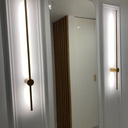 PROEFEKT Michał Czaplicki - Nowoczesne oświetlenie ścienne zintegrowane z panelami dekoracyjnymi, widoczne odbicie szafy z drewnianymi akcentami w lustrze, minimalistyczny design wnętrza.