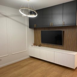 PROEFEKT Michał Czaplicki - Nowoczesny salon z drewnianą ścianą z lamelami, wiszącą szafką z czarnymi frontami i złotymi uchwytami, białą szafką RTV z ryflowanymi drzwiczkami oraz okrągłą lampą wiszącą z elementami w kolorze...
