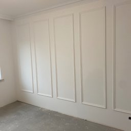 PROEFEKT Michał Czaplicki - Pokój z białymi ścianami ozdobionymi sztukaterią w formie prostokątnych paneli i listew przysufitowych, surowa podłoga w odcieniach szarości, widoczne gniazdko elektryczne.