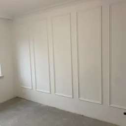 Pokój z białymi ścianami ozdobionymi sztukaterią w formie prostokątnych paneli i listew przysufitowych, surowa podłoga w odcieniach szarości, widoczne gniazdko elektryczne.