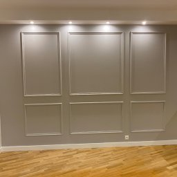 PROEFEKT Michał Czaplicki - Szara ściana z dekoracyjnymi listwami w kształcie paneli, drewniana podłoga, biała listwa przypodłogowa i fragment białej szafki po prawej stronie.