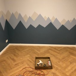PROEFEKT Michał Czaplicki - Pomieszczenie po świeżym pomalowaniu ściany w motyw górski, widoczne narzędzia malarskie w kartonie na parkiecie ułożonym w jodełkę.