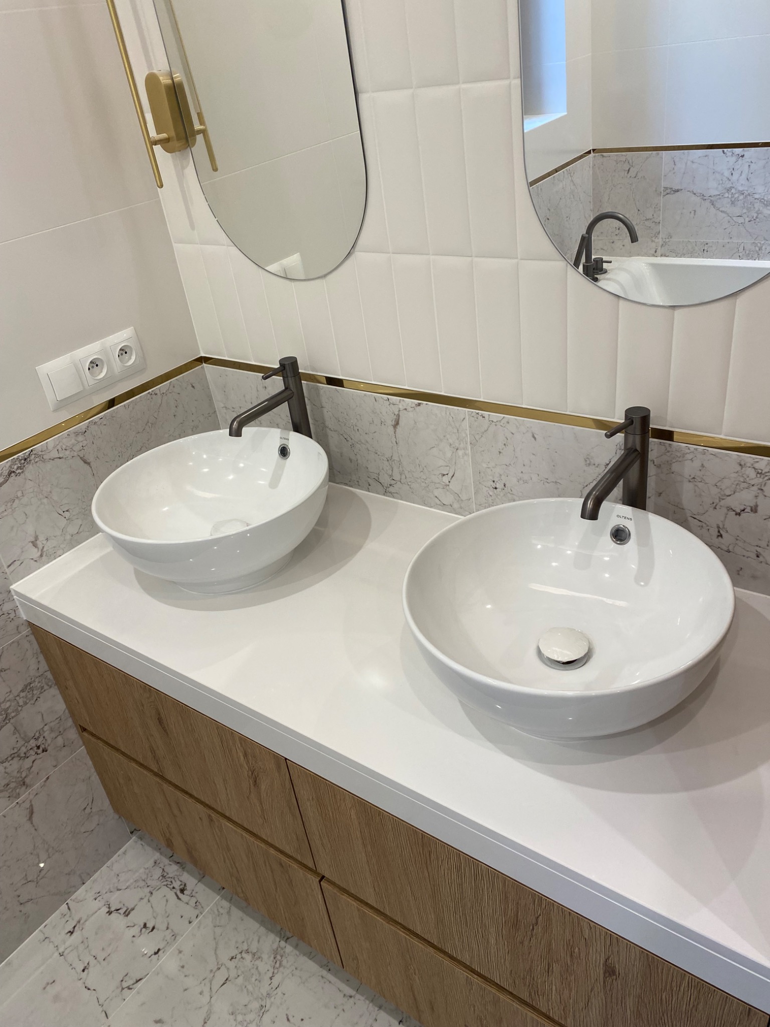 Nowoczesna łazienka z dwoma okrągłymi umywalkami nablatowymi, ciemnymi bateriami, marmurową ścianą z ozdobnym paskiem i owalnymi lustrami.