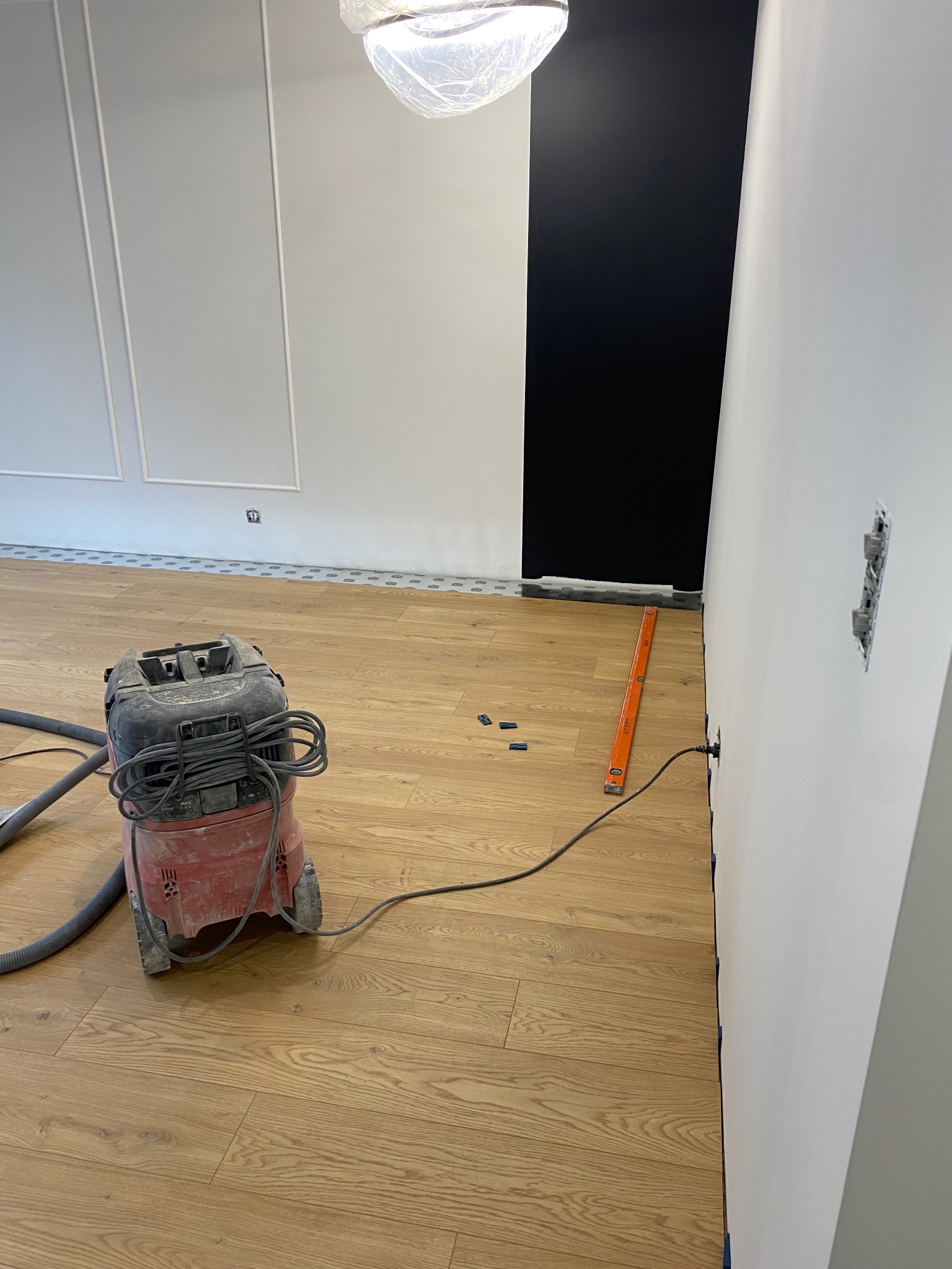 Podłoga z paneli po remoncie, widoczny odkurzacz przemysłowy, poziomica i listwy przypodłogowe zabezpieczone taśmą malarską, ściany białe z czarnym akcentem.